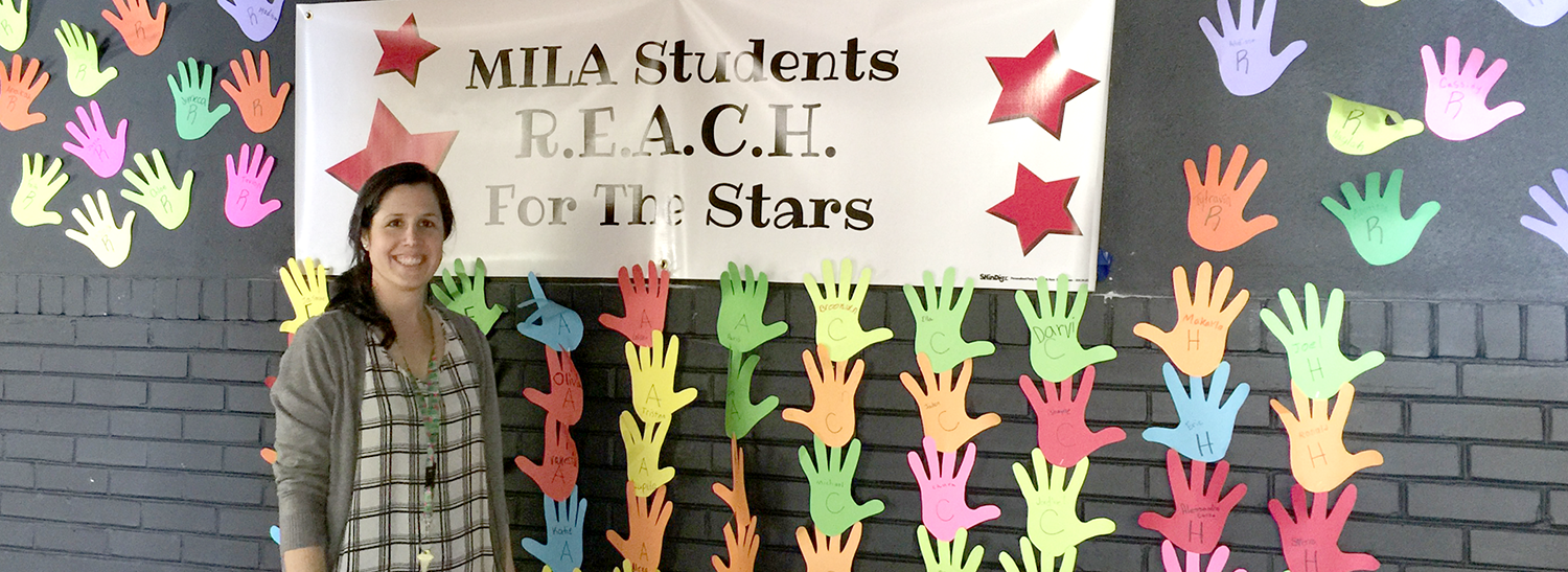 School teacher next to wall full of paper hands and sign that reads, 'MILA students R.E.A.C.H. for the stars.'